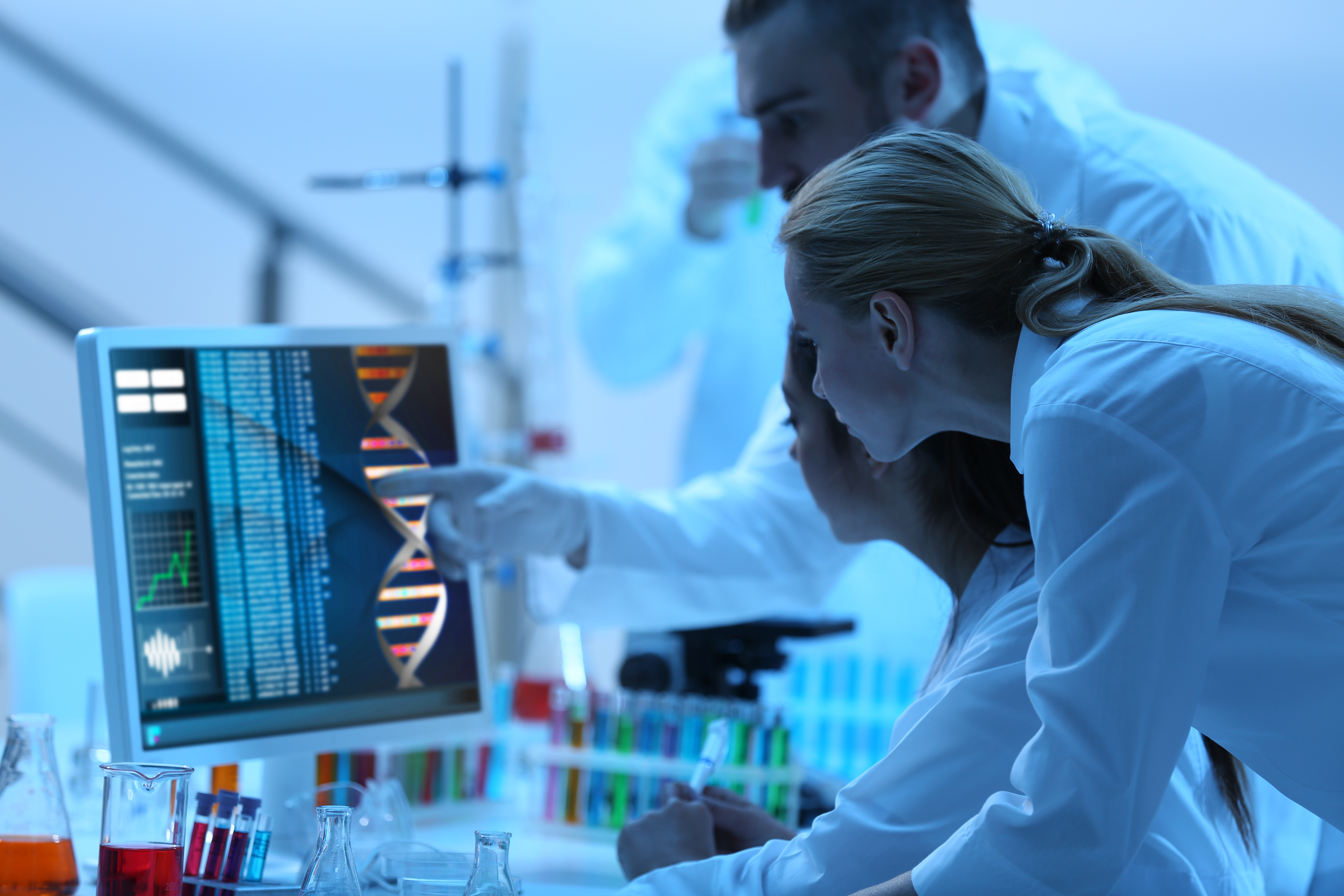 Três cientistas, vestidos de jaleco branco, olhando para a tela de computador, onde aparecem dados, represntando uma pesquisa clínica.
