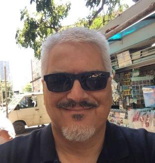 Homem branco de cabelos grisalhos, bigode e barba, vestindo camisa preta e usando óculos escuro
