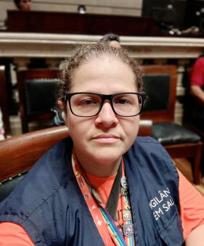 Mulher branca de cabelos castanhos, esticado e preso, usando óculos de armação preta, vestindo blusa laranja e colete azul sobre a blusa, com a descrição vigilância em saúde